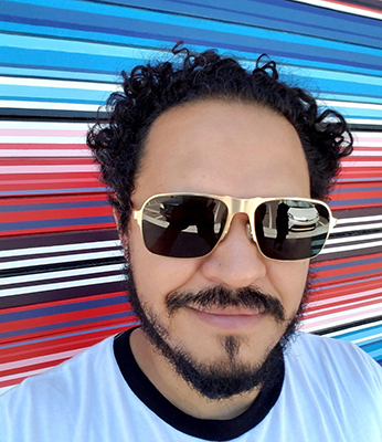 Latino man with curly black hair, sunglasses, and facial hair.